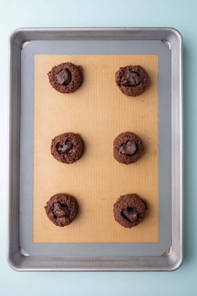 Frozen chocolate fudge sauce balls pressed into the center of chocolate cookie dough lined up on a baking sheet. 