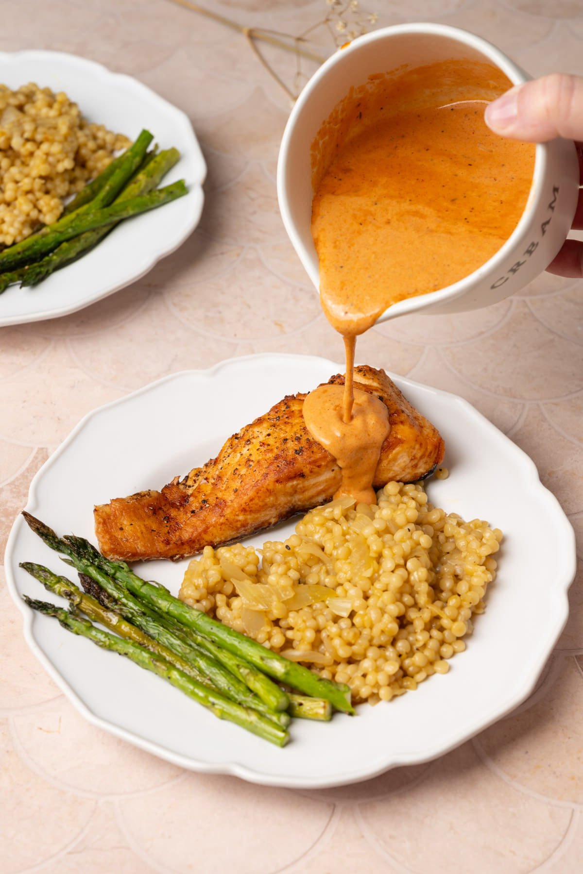 Pouring creamy Cajun sauce over salmon on a dinner plate with couscous and asparagus. 