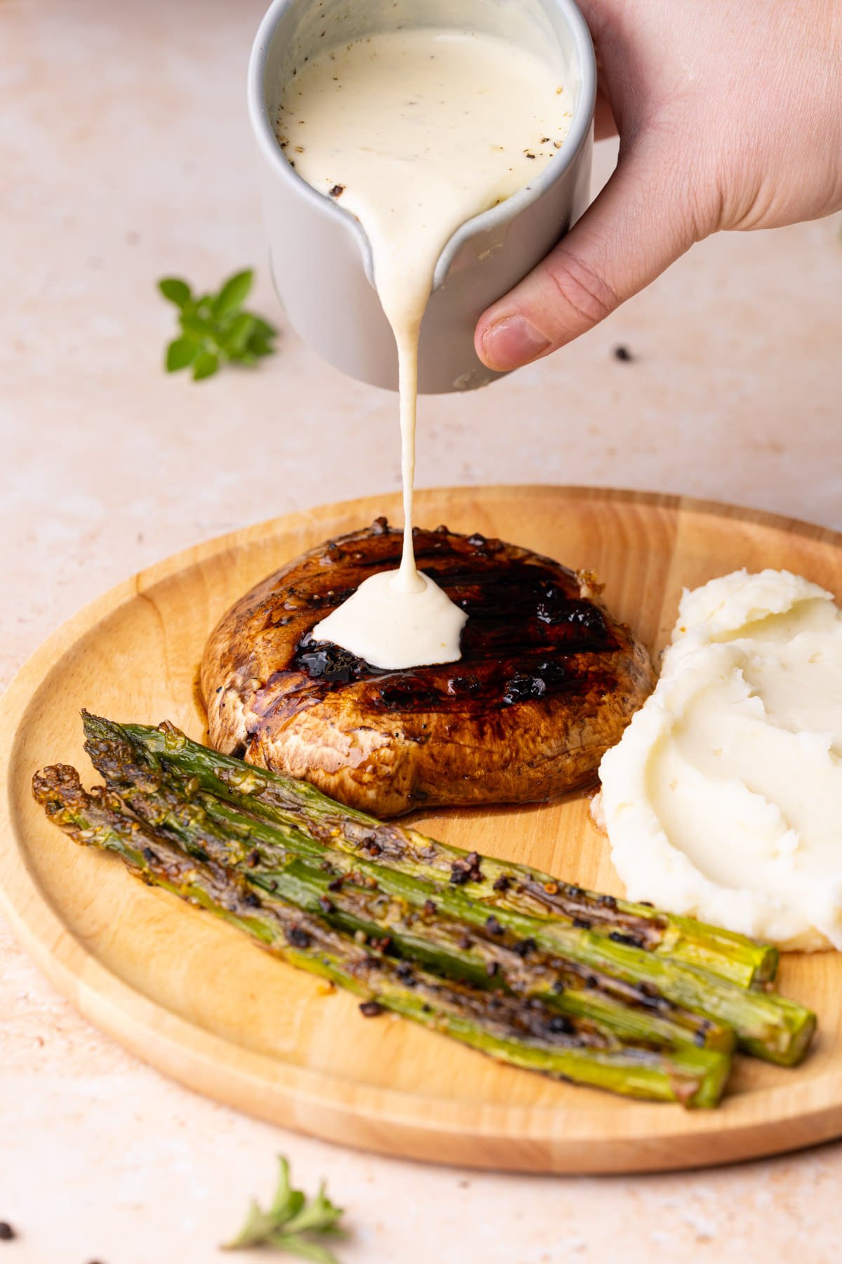 Pouring creamy Parmesan garlic sauce over dinner plate with portobello mushroom, asparagus, and mashed potato. 