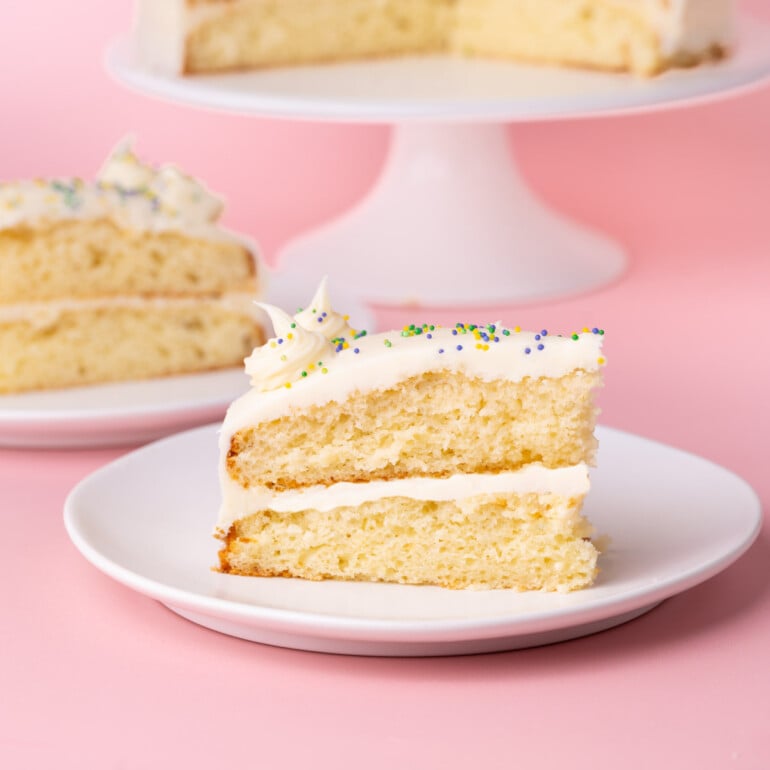 Slice of layered white cake on a plate with white chocolate ganache and colorful sprinkles, with the whole cake in the background.