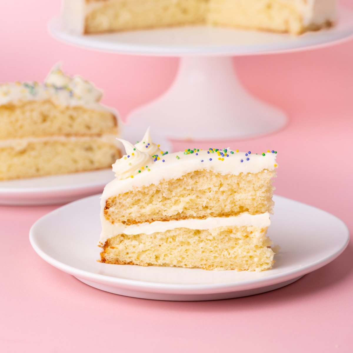 Slice of layered white cake on a plate with white chocolate ganache and colorful sprinkles, with the whole cake in the background.