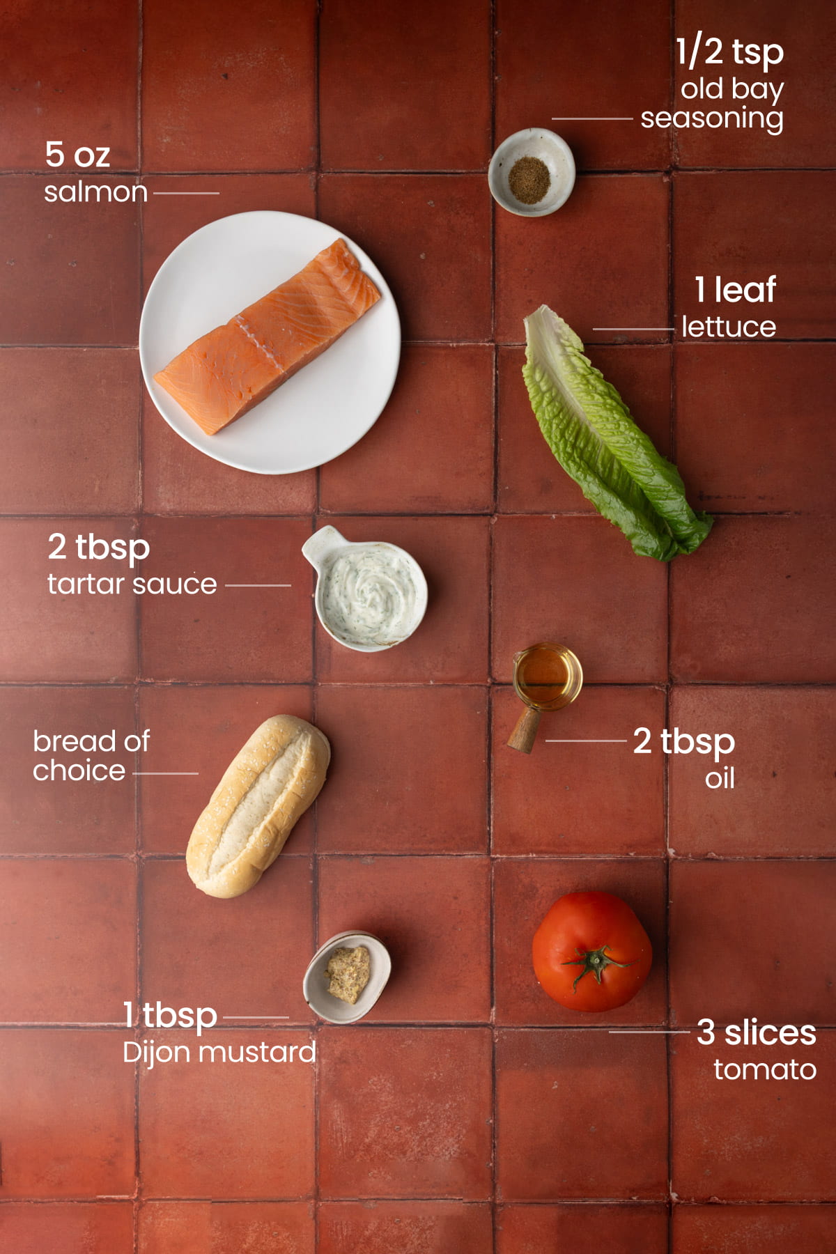 Overhead view of all the ingredients needed for a pan-fried salmon sandwich - old bay seasoning, lettuce, salmon, tartar sauce, oil, bread, Dijon mustard, and tomato.