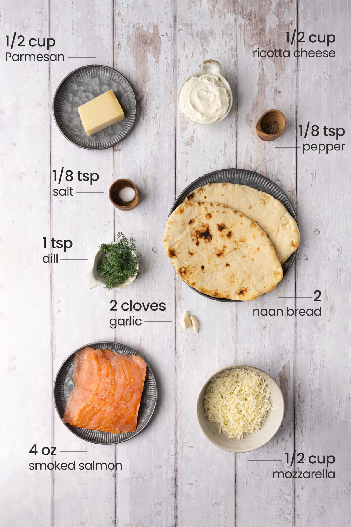 Overhead view of the ingredients for smoked salmon flatbread - Parmesan, ricotta cheese, salt, pepper, dill, garlic, naan bread, smoked salmon, and mozzarella.