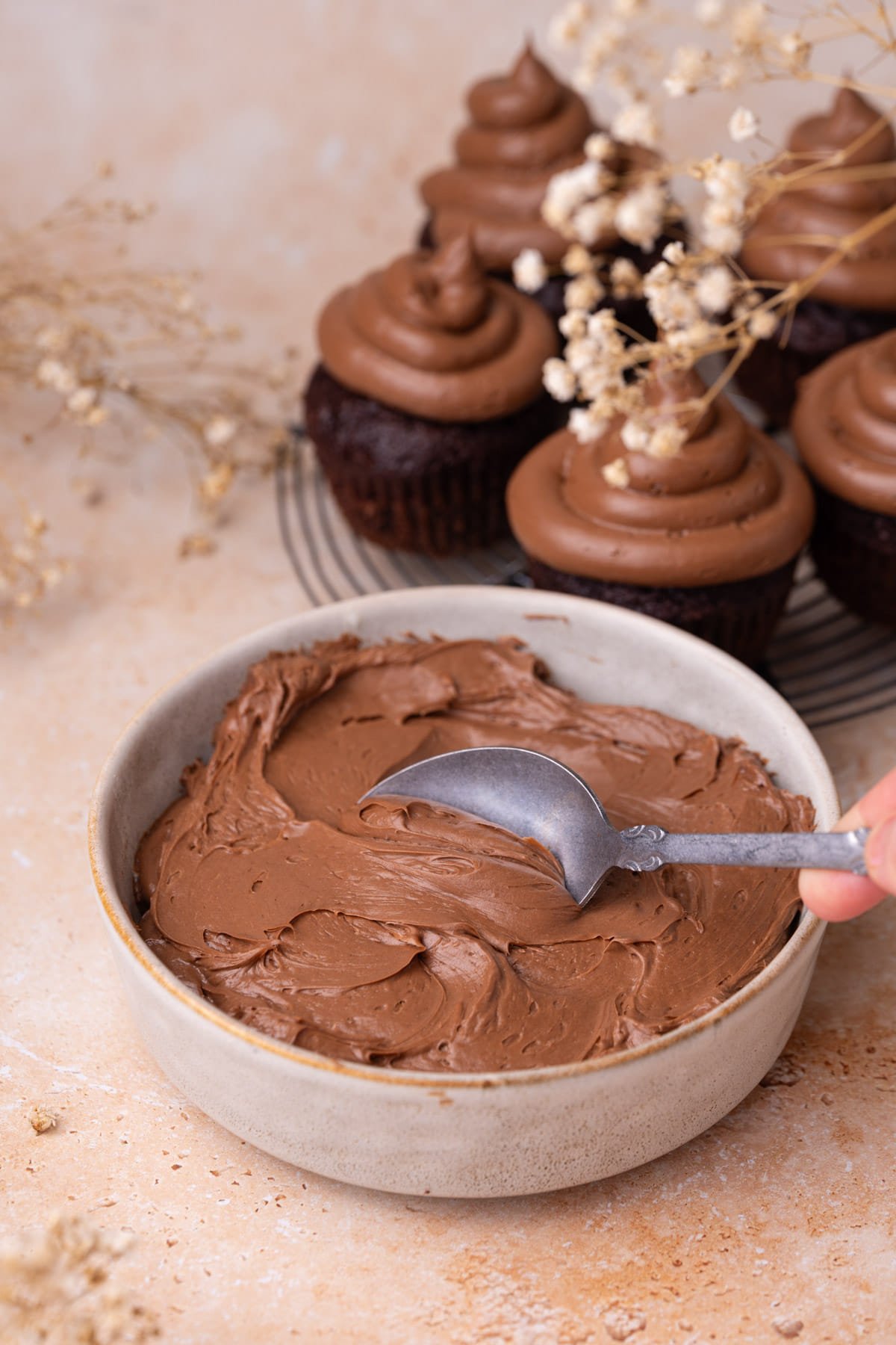 Cream cheese chocolate frosting in a bowl with frosted cupcakes in background. 