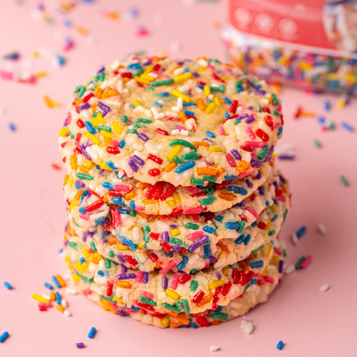 Stack of sprinkle sugar cookies with jar of rainbow sprinkles in the background.