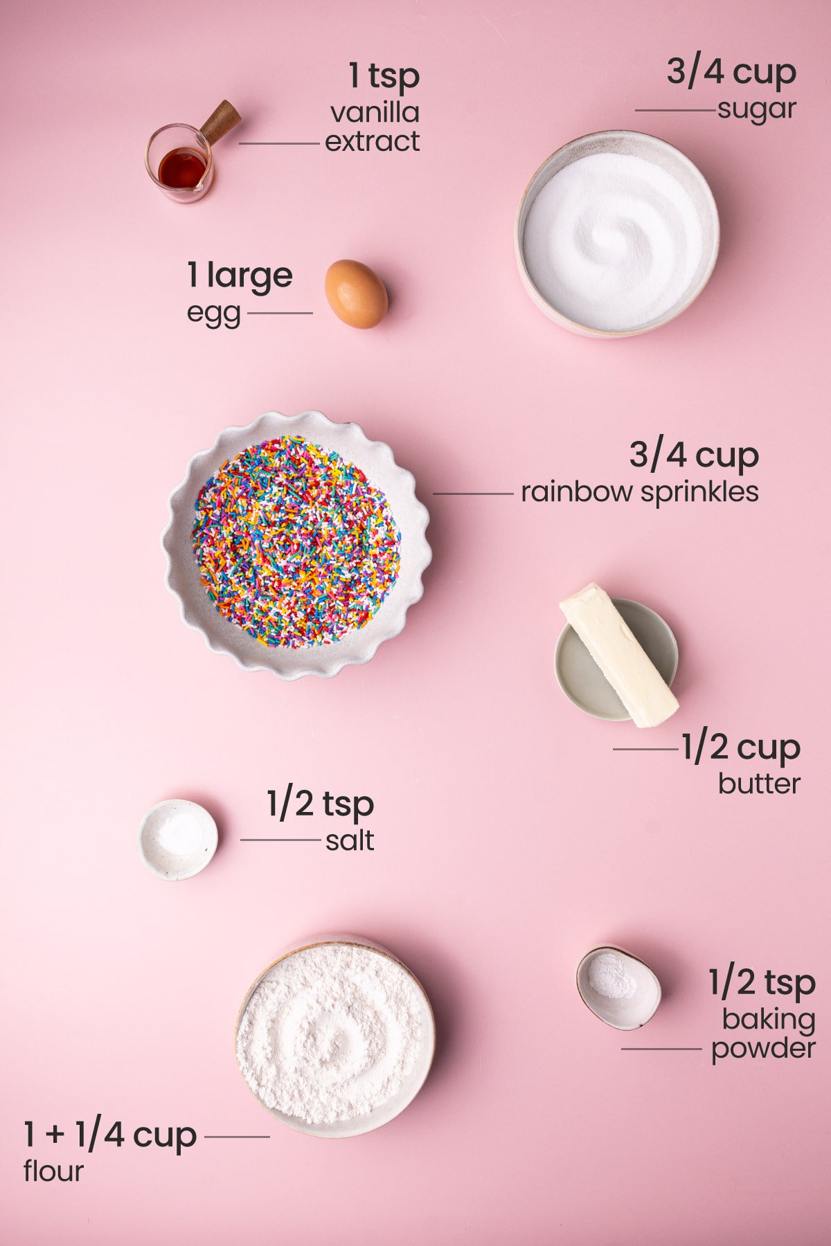 Overhead view of the ingredients needed for sprinkle sugar cookies - vanilla extract, sugar, egg, rainbow sprinkles, salt, butter, flour, and baking powder.