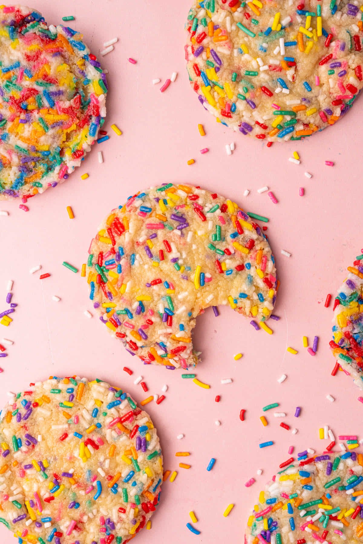 Overhead image of sugar cookies with rainbow sprinkles. middle cookie has a bite taken out of it.