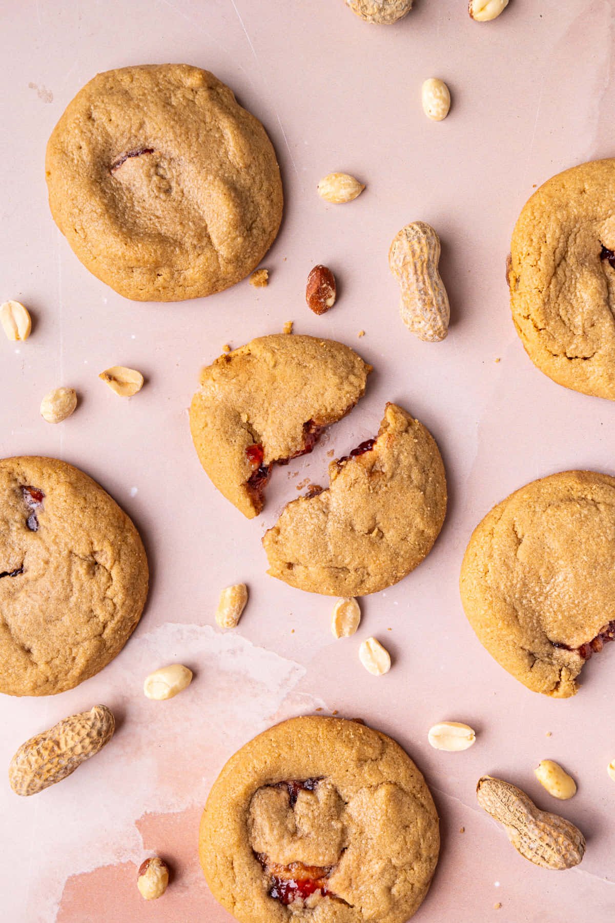 Overhead image of peanut butter and jelly cookies on a pink surface with the centermost one broken in half. 