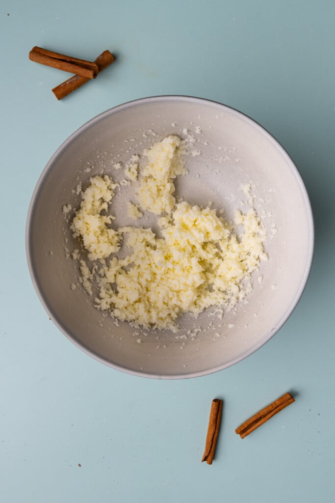 Creaming butter and granulated sugar together in a large mixing bowl with cinnamon sticks in the background. 