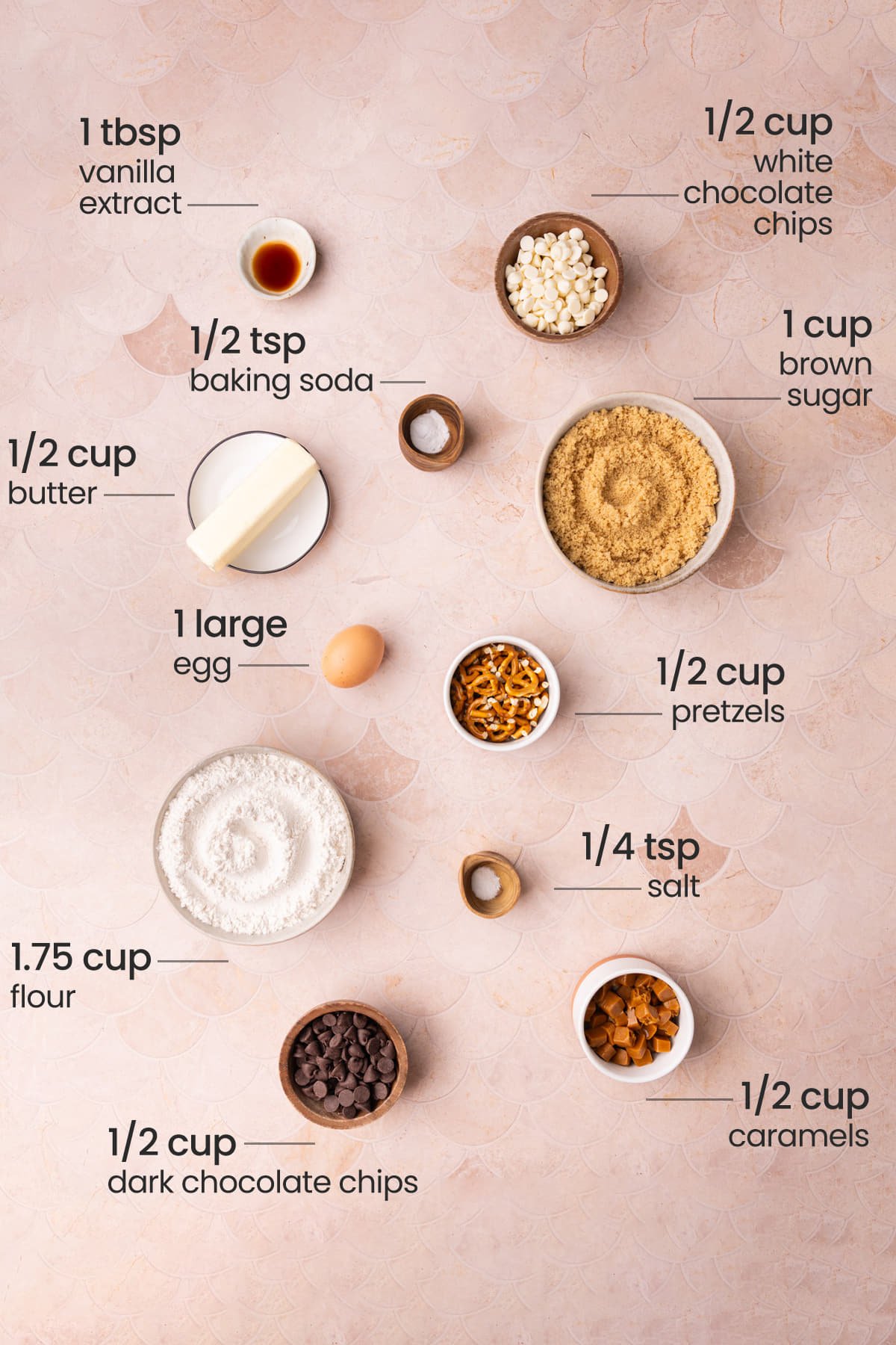 All ingredients needed for Kitchen Sink Cookies including vanilla extract, white chocolate chips, baking soda, butter, brown sugar, egg, pretzels, flour, salt, dark chocolate chips, and caramels. 
