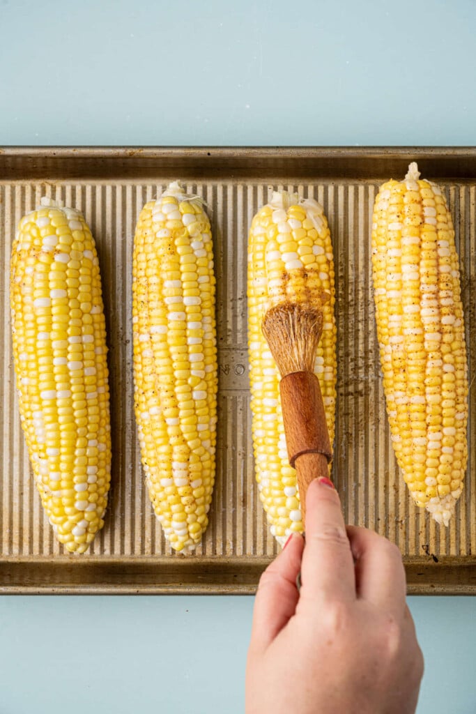 Brushing Cajun butter onto corn on the cob before roasting it in the oven.