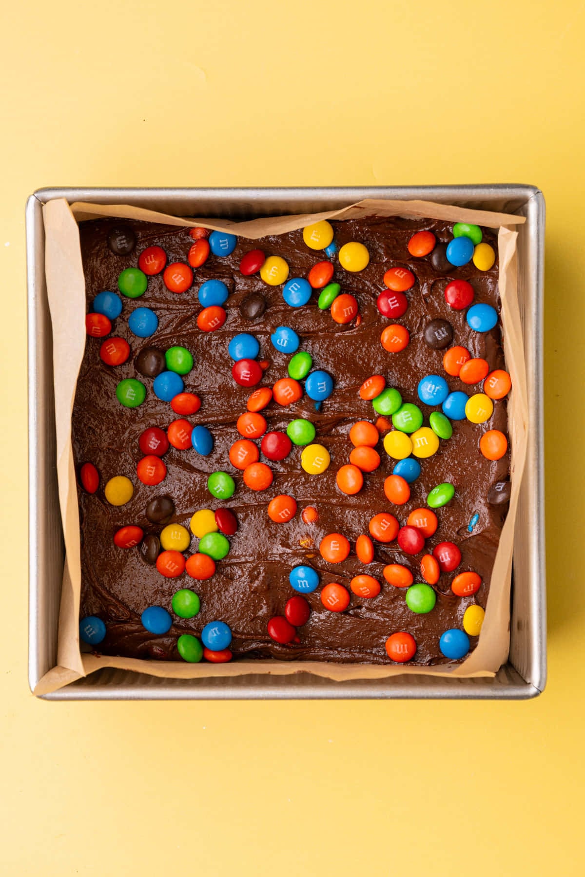 Brownie batter spread evenly in a lined square baking pan with M&M's sprinkled on top. 