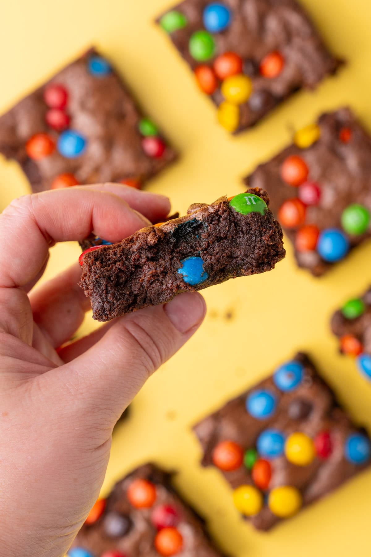 A batch of M&M brownies on a yellow background, holding the center brownie up to the camera to see the side view. 