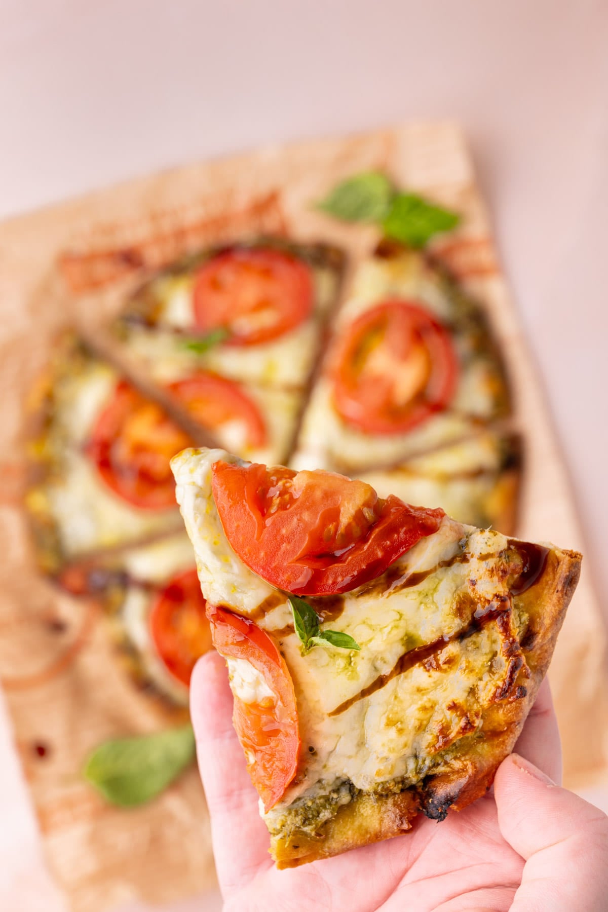 Holding a slice of naan pizza with pesto to the camera to show the melted cheese, fresh tomatoes, and balsamic glaze.