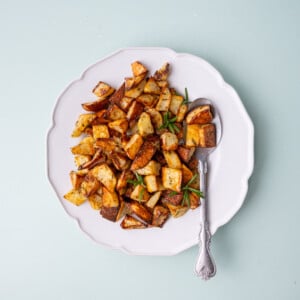 Crispy breakfast potatoes with rosemary on a white scalloped plate with a serving spoon.