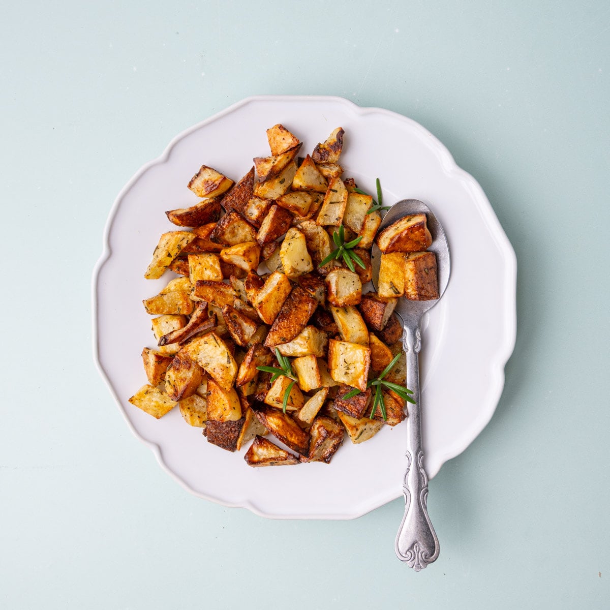 Crispy breakfast potatoes with rosemary on a white scalloped plate with a serving spoon.