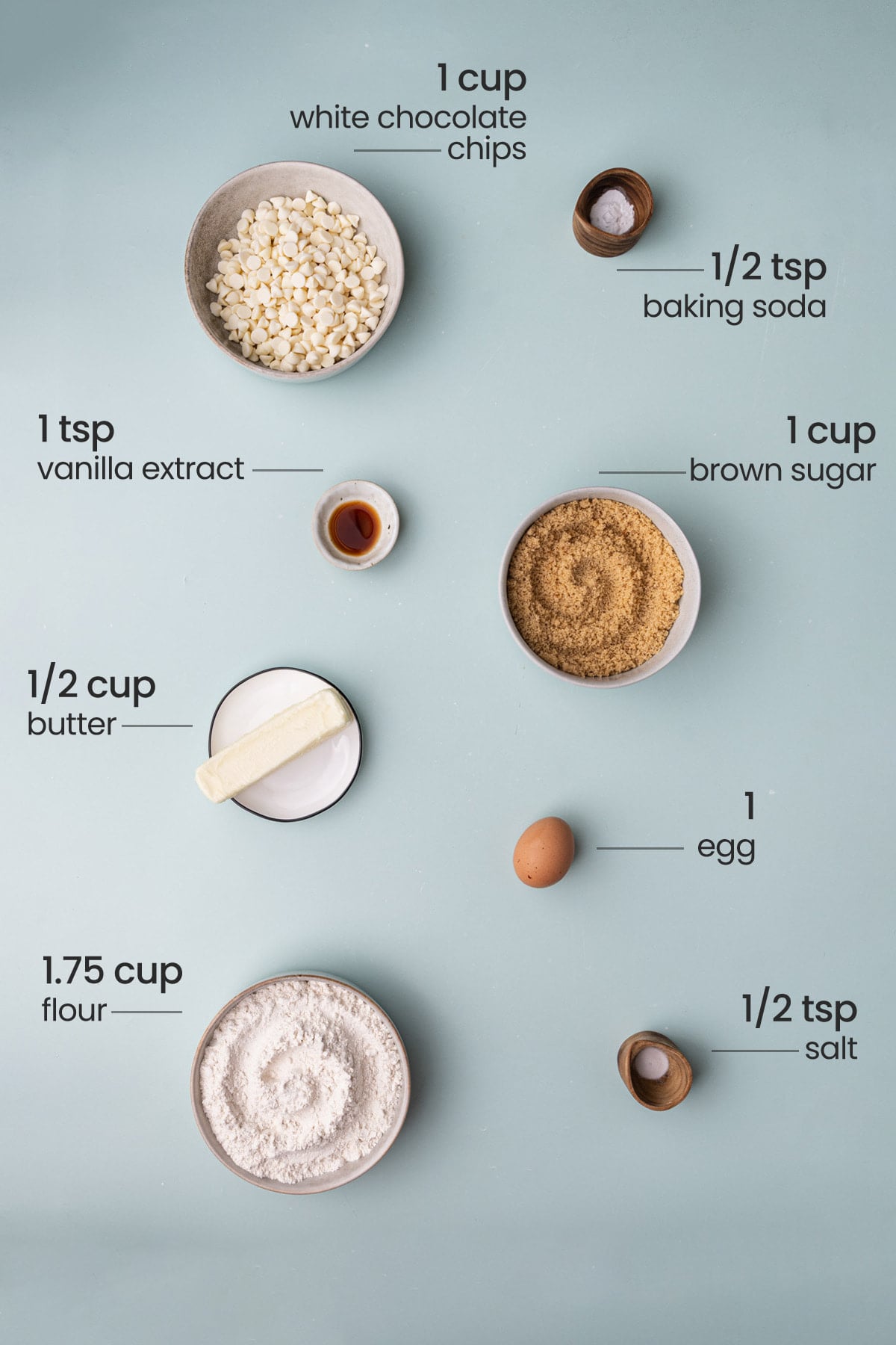 All ingredients required for white chocolate chip cookies, including baking soda, white chocolate chips, vanilla extract, light brown sugar, butter, egg, all-purpose flour, and salt.