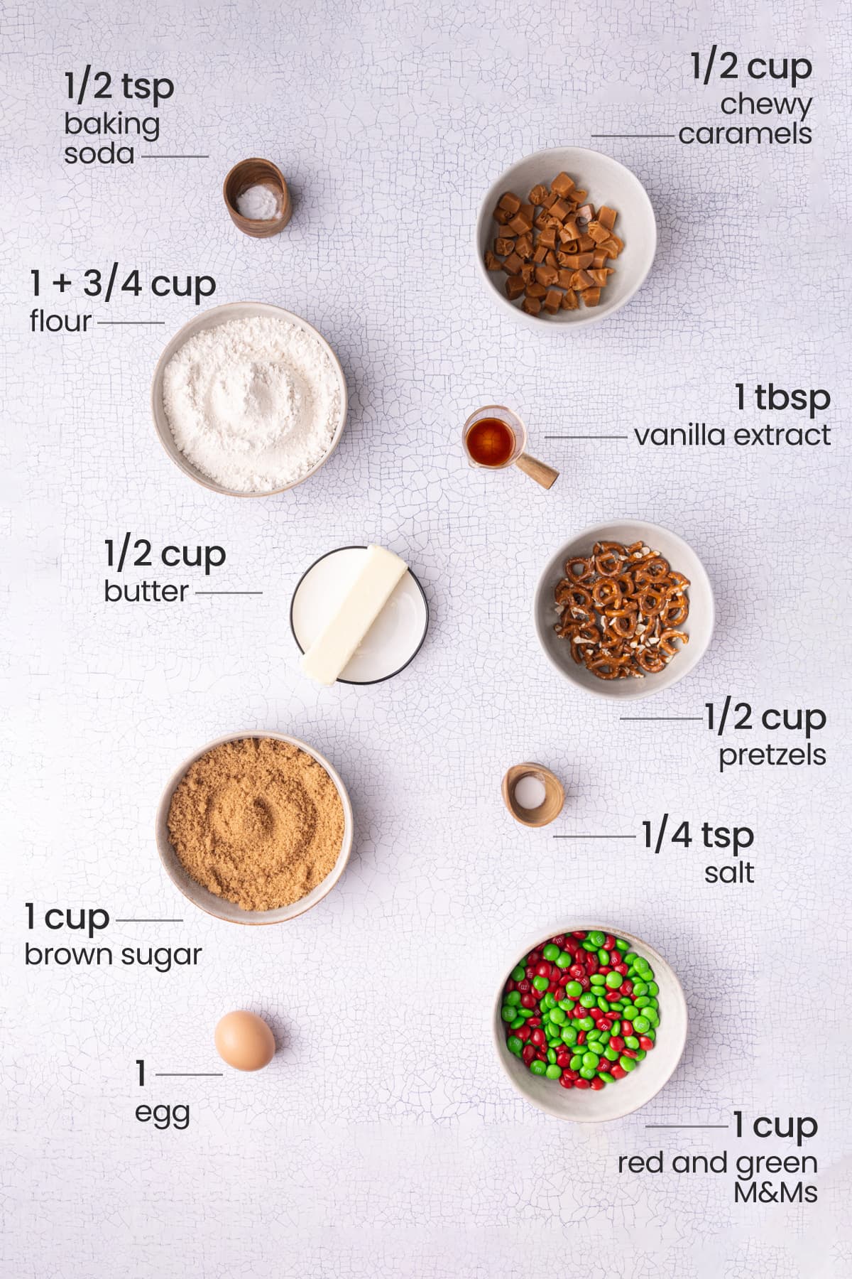 Overhead view of the ingredients for Christmas kitchen sink cookies - baking soda, chewy caramels, flour, vanilla extract, butter, pretzels, brown sugar, salt, egg, and red and green M&Ms.