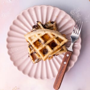 Overhead image of chocolate chip waffles cut into quarters and stacked on a pink scalloped plate with syrup.