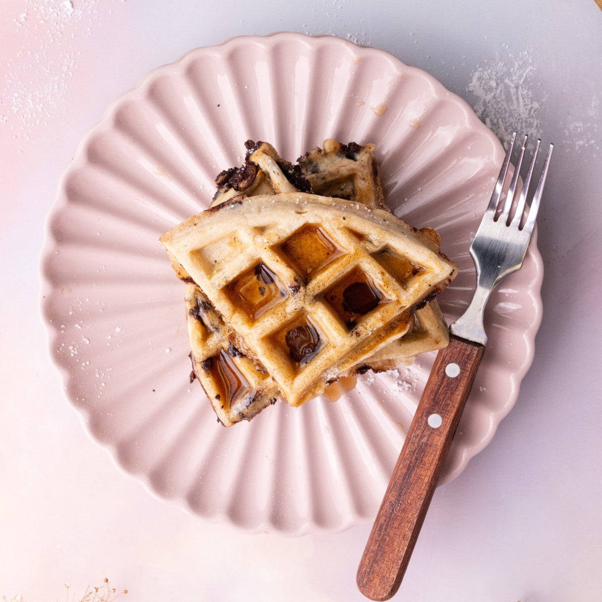 Overhead image of chocolate chip waffles cut into quarters and stacked on a pink scalloped plate with syrup.