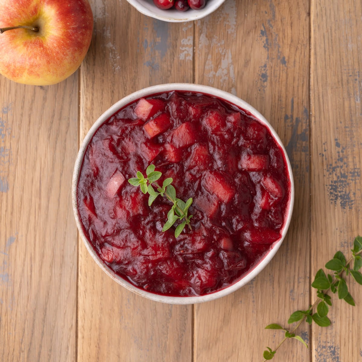 Overhead image of a bowl of vibrant homemade cranberry sauce with apples garnished with fresh thyme.