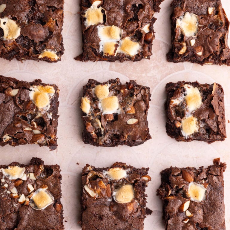 Batch of Rocky Road brownies with toasted marshmallows and almonds on top lined up on a peach tile background.