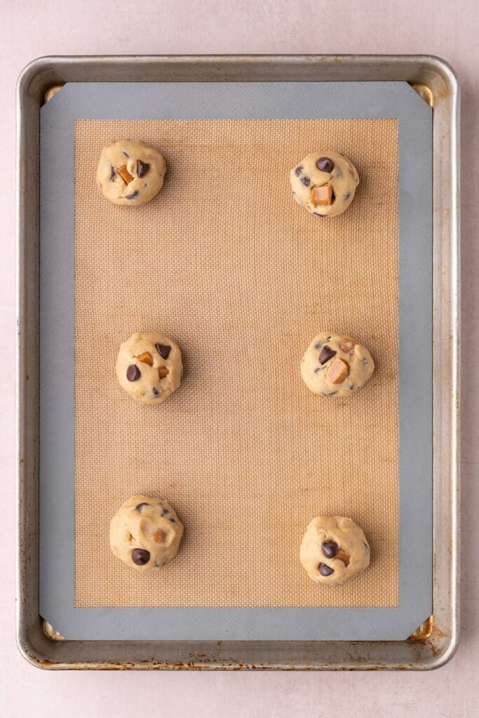 Cookie dough formed into round cookies with chocolate chips and caramels on a lined baking sheet ready for the oven.