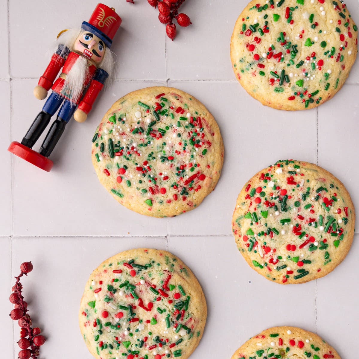 Christmas Sprinkle Cookies Featured Image Christmas sugar cookies with red and green sprinkles on white tile with a little nutcracker Christmas figurine next to them.