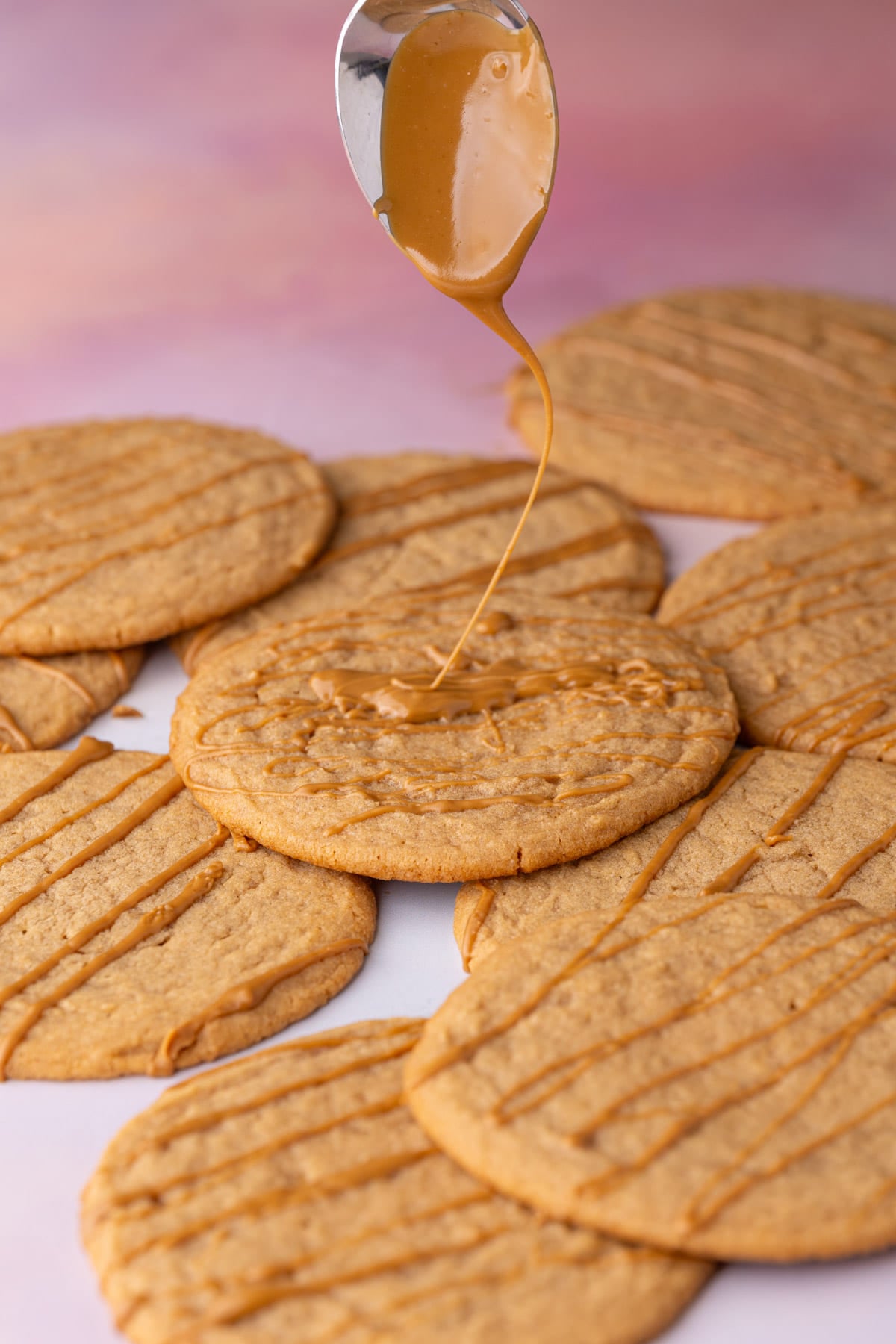 Drizzling extra cookie butter over thin and chewy Biscoff cookie butter cookies on a colorful surface with a spoon.