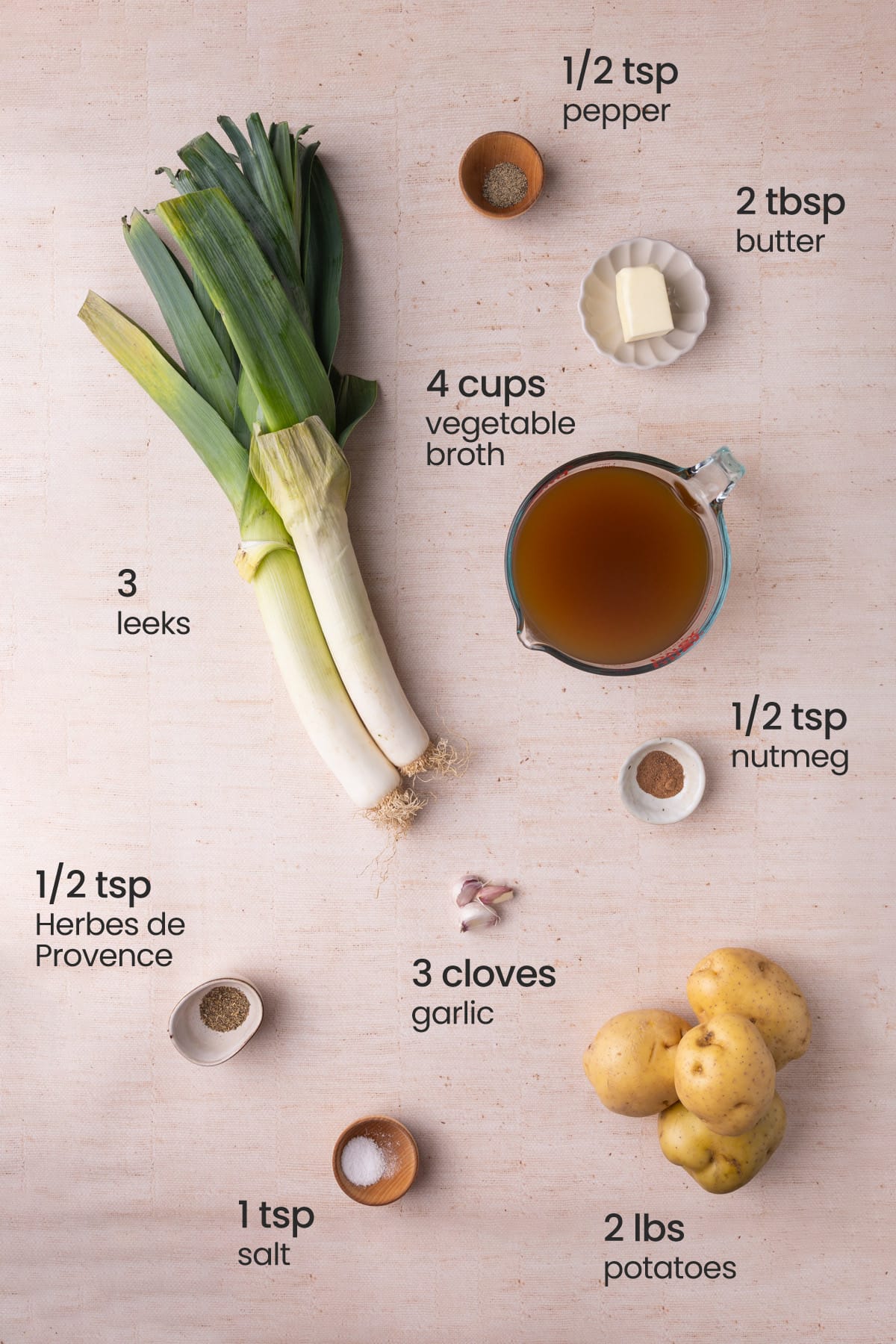 Overhead view of the ingredients needed for potato and leek soup - pepper, butter, vegetable broth, leeks, nutmeg, garlic, salt, potatoes and Herbes de Provence.