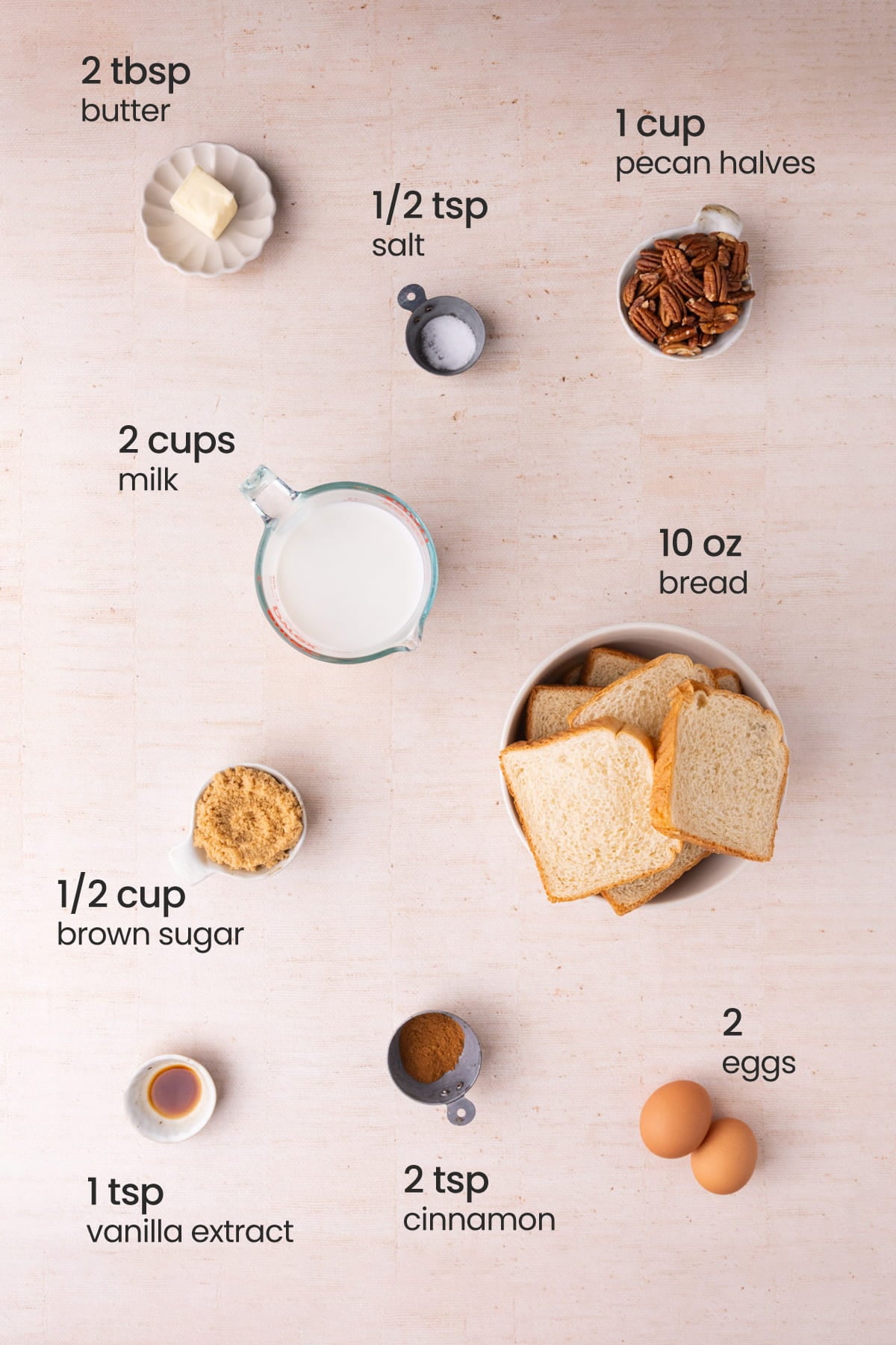 Overhead view of the ingredients needed for pecan pie bread pudding - butter, salt, pecan halves, milk, bread, brown sugar, vanilla extract, cinnamon, and eggs.