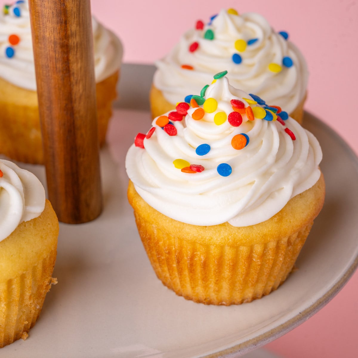 Close up of a vanilla cupcake with a generous amount of sour cream frosting finished with rainbow sprinkles on a cupcake display.