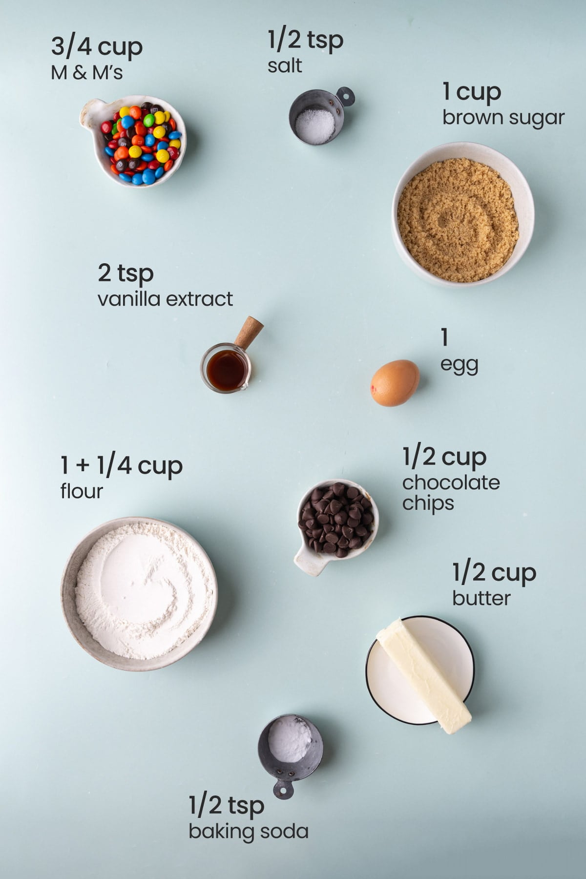 Overhead view of the ingredients needed for One-Bowl Chewy M&M’s Cookie Bars - M&M's, salt, brown sugar, vanilla extract, egg, flour, chocolate chips, butter, and baking soda.