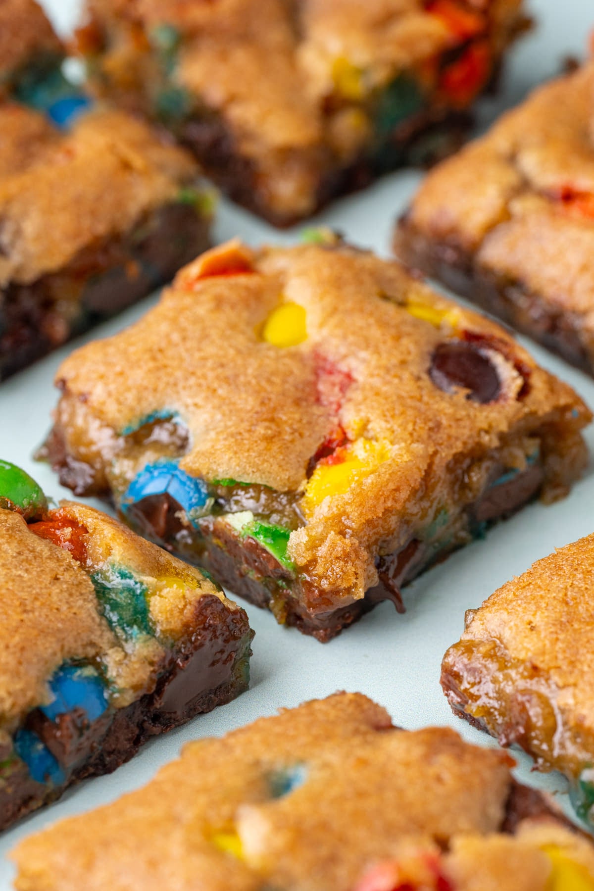 Chewy cookie bars with M&M's sliced into squares on a blue background with picture taken at an angle to show the textures of both the top and inside.