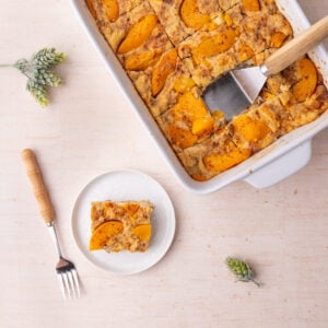 Peach bread pudding still in a casserole dish with a slice scooped out and served on a small white plate.