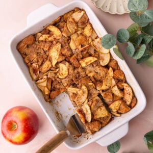 Baking dish with freshly baked homemade bread pudding with apples, with two slices missing and a fresh apple on the side.