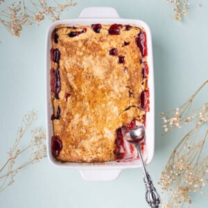 Cherry Cheesecake dump cake with a serving spoon, showing layers of buttery yellow cake mix, juicy fruit filling, and dollops of cream cheese.